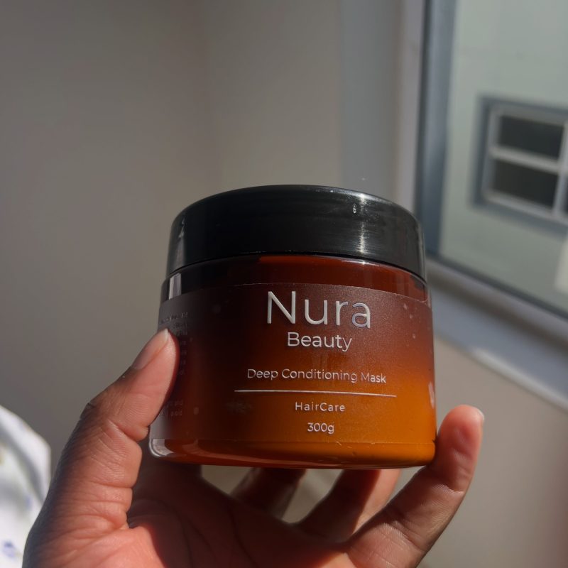 A hand holding the Nura Beauty Deep Conditioning Mask, a 300g amber jar, in warm daylight