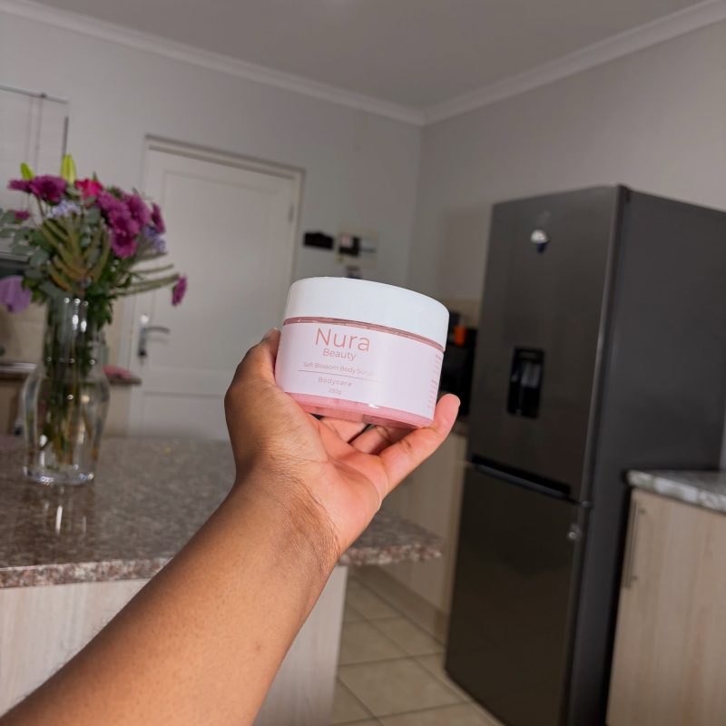 A hand holding the Nura Beauty Soft Blossom Body Scrub jar, with fresh flowers behind